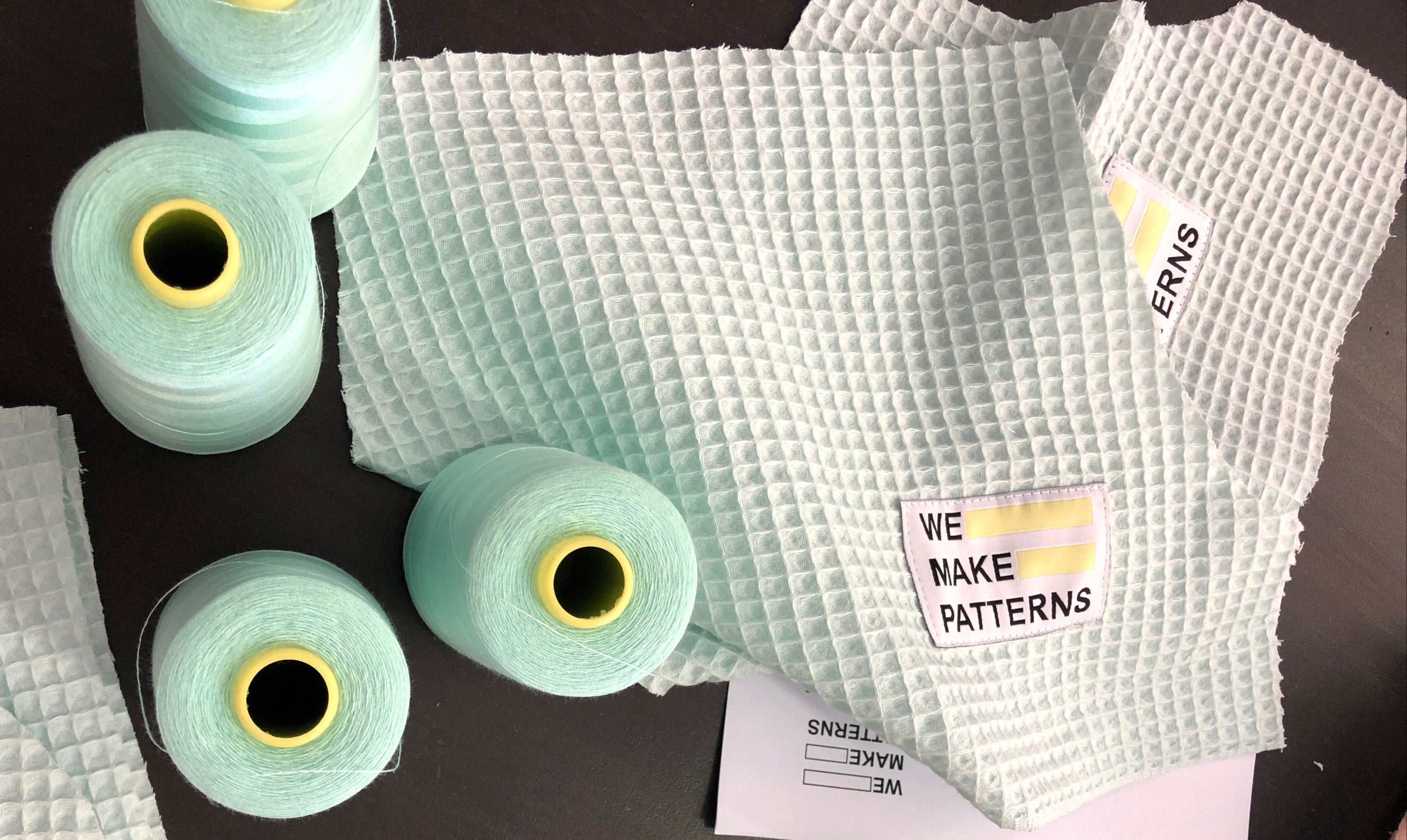 Stoffzuschnitte und Garnrollen in der Farbe Mint liegen auf einem schwarzen Tisch Mit angenähten We Make Patterns Labels